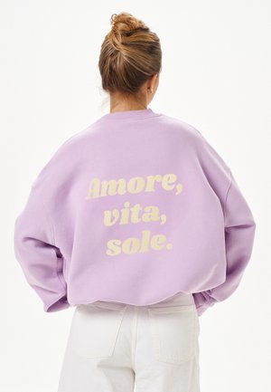 Person mit blondem Haar im Dutt, die ein hellviolettes Sweatshirt mit dem Schriftzug „Amore, vita, sole.“ auf dem Rücken und weiße Hosen trägt.