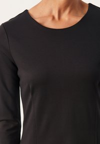 Haut noir à manches longues avec un col rond et un tissu lisse et extensible. Coupe ajustée avec des détails discrets de coutures le long des côtés.