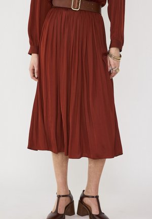 Roestkleurige geplooide midi-rok met bruine leren riem, gecombineerd met bruine T-strap blockhakschoenen en gouden armbanden en ringen aan één hand.