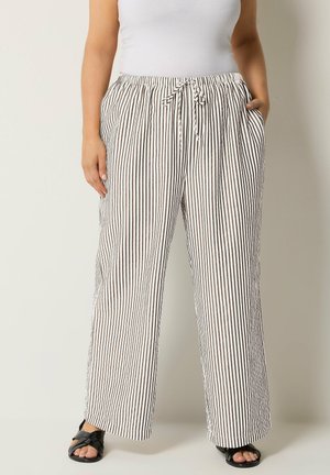 Person trägt lockere, hochtaillierte weiße Hosen mit dünnen schwarzen vertikalen Streifen und einem Kordelzug, kombiniert mit schwarzen Sandalen.