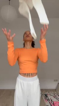 Persona oranžā garā krekla un baltu biksu tērpā met baltu apģērbu gaisā telpā ar paklāju un pakaramo gaismekli.