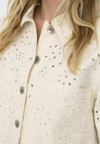 Veste courte couleur crème en tissu brodé, dotée d'une fermeture à boutons, de deux poches poitrine et d'un motif floral dans le matériau.