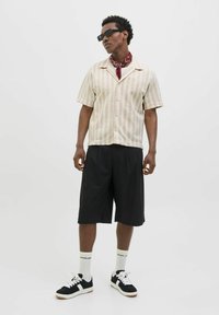 Chemise à manches courtes rayée en beige et blanc, associée à un short plissé noir. Les accessoires comprennent un bandana rouge et des lunettes de soleil noires.