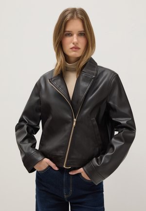 Junge Frau trägt eine schwarze Lederjacke über einem beigen Rollkragenpullover und steht mit den Händen in den Taschen ihrer dunkelblauen Jeans vor einem einfarbigen Hintergrund.