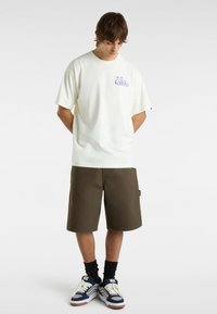 Camiseta blanca de algodón con el logo morado de Vans, combinada con pantalones cortos marrones oscuros y zapatillas azules. El modelo está de pie con la cabeza agachada y las manos detrás de la espalda.