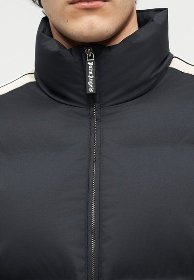 Nærbillede af en person iført en sort polstret jakke med høj krave og en lynlåstrækker mærket "Palm Angels".