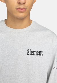 Nahaufnahme einer Person, die einen hellgrauen Sweatshirt mit dem schwarzen Text "Element" auf der linken Brust trägt.