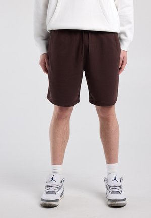 Man draagt donkerbruine shorts, witte sokken, witte en grijze sneakers en een wit hoodie, staand tegen een egale achtergrond.