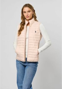 Lichtroze gewatteerde vest met verticale gestikte ontwerpen, hoge kraag en marineblauw logo. Draagt over een witte geribde lange mouw top en blauwe jeans.