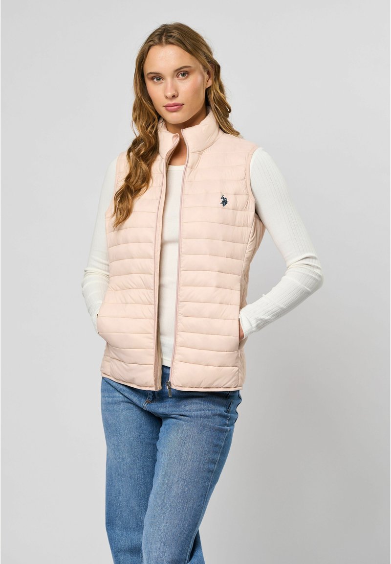 Lichtroze gewatteerde vest met verticale gestikte ontwerpen, hoge kraag en marineblauw logo. Draagt over een witte geribde lange mouw top en blauwe jeans.