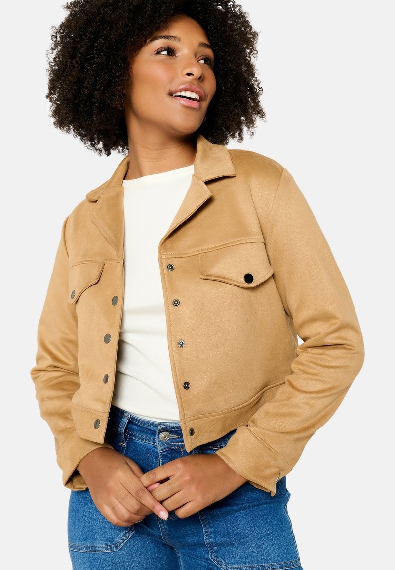 Beige Wildleder-Cropped-Jacke mit Knopfleiste, zwei Brusttaschen und Kragen, getragen über einem weißen Oberteil und blauen Jeans.