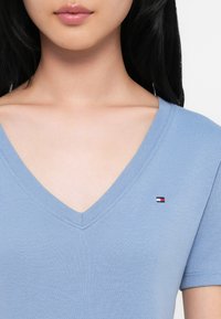 Lyseblå V-hals t-skjorte, korte ermer, glatt tekstur, med en liten logo på øvre venstre side.