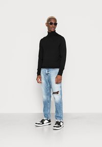 Maglione nero a collo alto, jeans azzurri chiari strappati con tagli e sneaker bianco e nere. Gli accessori includono occhiali da sole scuri.