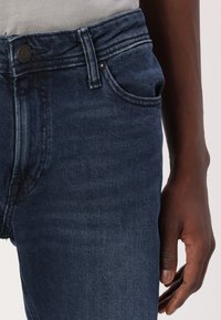 Dark blue denim jeans with a classic five-pocket design, a buttoned fly, and subtle stitching details along the waistband and pockets.