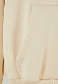 Beige huppari, jossa on kengurupocketti, pehmeä rakenne, ribattu helma ja näyttävä tikkaus.