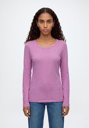 Femme aux longs cheveux bouclés portant une chemise à manches longues violet clair et un jean bleu, debout devant un fond blanc uni.