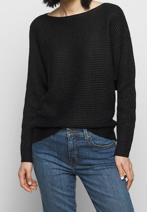 Person trägt einen schwarzen, gerippten, langärmligen Pullover aus Strick, der in eine mittelhohe blaue Jeans mit Knopf- und Reißverschluss gesteckt ist.