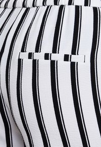 Tissu à rayures noir et blanc, présentant des lignes verticales de largeurs variées. Comprend une petite poche avec un contour cousu subtil.