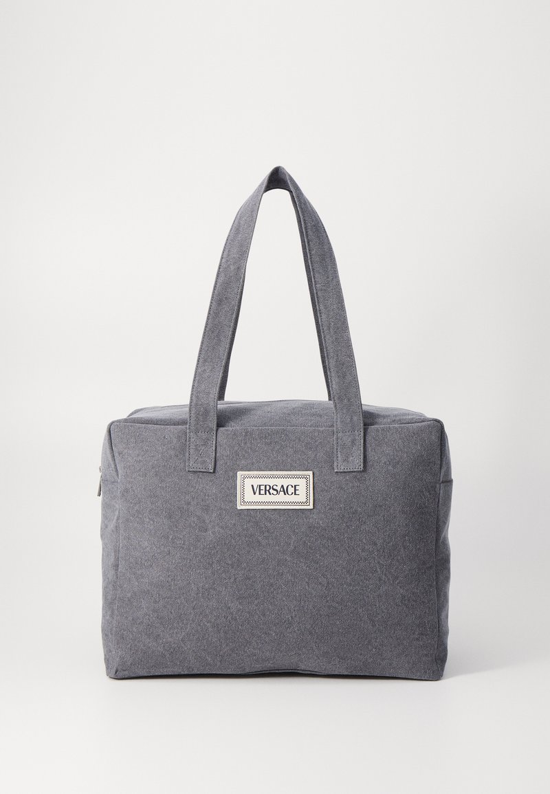 Bolsa de lona cinza com duas alças robustas, fecho de correr e um distintivo de logotipo em destaque na frente. Forma retangular com uma superfície texturizada.