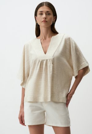 Top beige largo con scollatura a V, maniche corte e dettagli plissettati. Abbinato a pantaloni corti bianchi. Tessuto leggero e testurizzato.