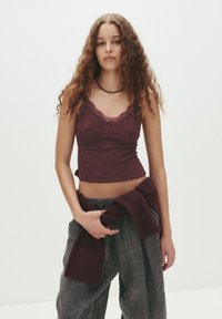 Burgundy croppad topp med texturerad tyg och spetsdetaljer, kombinerad med lösa grå byxor och en matchande stickad tröja knuten vid midjan.