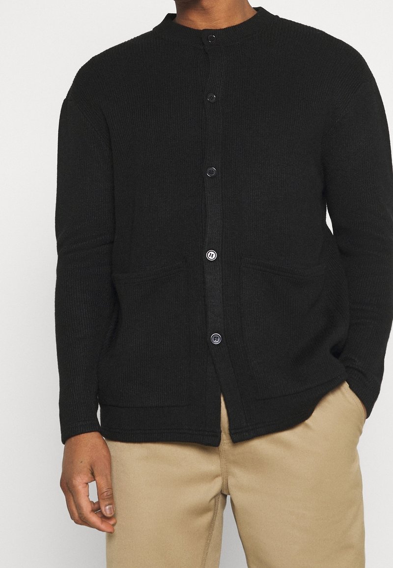 Homme portant un cardigan noir boutonné avec deux poches avant et un pantalon beige, debout avec une main dans la poche et l'autre détendue.