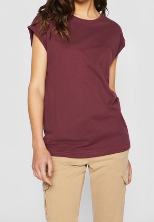 Kvinde iført en kortærmet mørkerød T-shirt og beige cargobukser, vist fra skuldre til hofter mod en ensfarvet baggrund.
