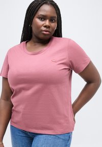 Γυναίκα με μακριά κοτσίδα, φορώντας ένα ροζ μπλουζάκι Levi's και μπλε τζιν, στέκεται με το ένα χέρι στη μέση της, πάνω σε λευκό φόντο.