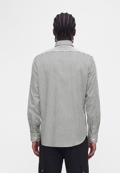 Homme aux cheveux tressés portant une chemise à rayures verticales noires et blanches avec des boutons et un pantalon noir, vu de dos sur un fond blanc.