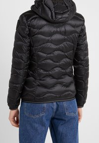 Schwarze Pufferjacke mit Kapuze, gequiltetem Design, glänzender Textur und elastischen Bündchen. Getragen über einer Paar blauer Jeans.