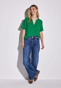 Blusa verde a maniche corte con scollo a V, abbinata a jeans blu a gamba larga e ballerine marroni con stampa leopardata. Tessuto morbido, vestibilità casual.