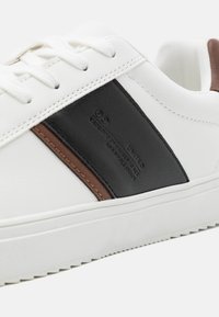 Weiße Sneakers mit schwarzen und braunen Lederakzenten, ausgestattet mit strukturierten Materialien, flachen Schnürsenkeln und einer strukturierten Gummisohle.