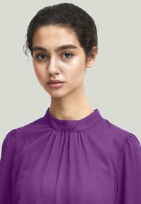 Blusa viola con colletto alto e dettagli arricciati. Tessuto morbido e leggero splendore, con maniche a sbuffo per un volume aggiunto.