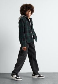 Plaid flannel jacket in black and green, gray hoodie underneath, paired with dark pants and black/white sneakers. Smooth textures, relaxed fit.