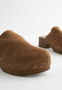 Bruine suède klompen met een ronde neus, houten platform en zichtbare metalen noppen langs de buitenzoel, met een gladde structuur.