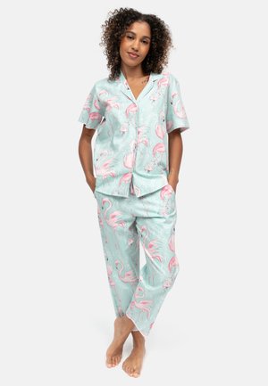 Mujer con cabello rizado que lleva un pijama azul claro con estampado de flamencos rosas, de pie descalza con las manos en los bolsillos contra un fondo blanco.