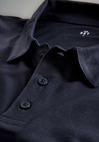 Temno modra polo majica z mehko teksturo, ki ima klasičen ovratnik, dva črna gumba in subtilne šiviljske detajle.