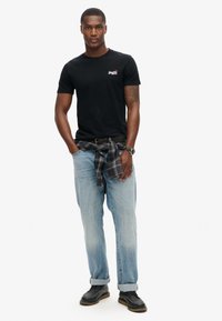 T-shirt nera in cotone con logo, jeans in denim sbiadito blu chiaro, scarpe nere e camicia a quadri annodata in vita.