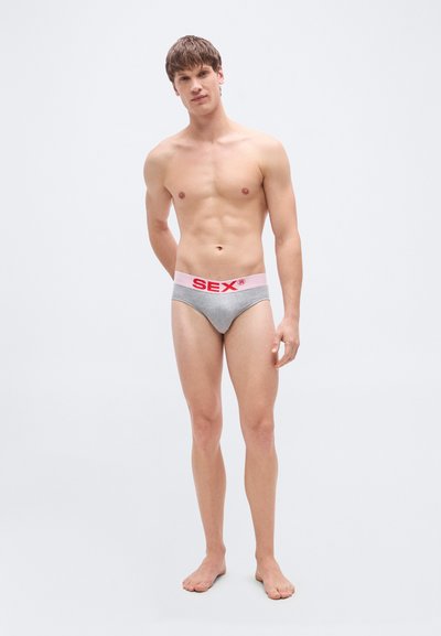 Sive bombažne spodnjice z roza pasom, na katerem je v debelih rdečih črkah napis "SEX". Model stoji bosa.
