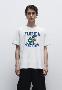Witte katoenen t-shirt met de tekst "FLORIDA GATORS" in oranje en blauw, met een groene alligator graphic. Ontspannen pasvorm en korte mouwen.