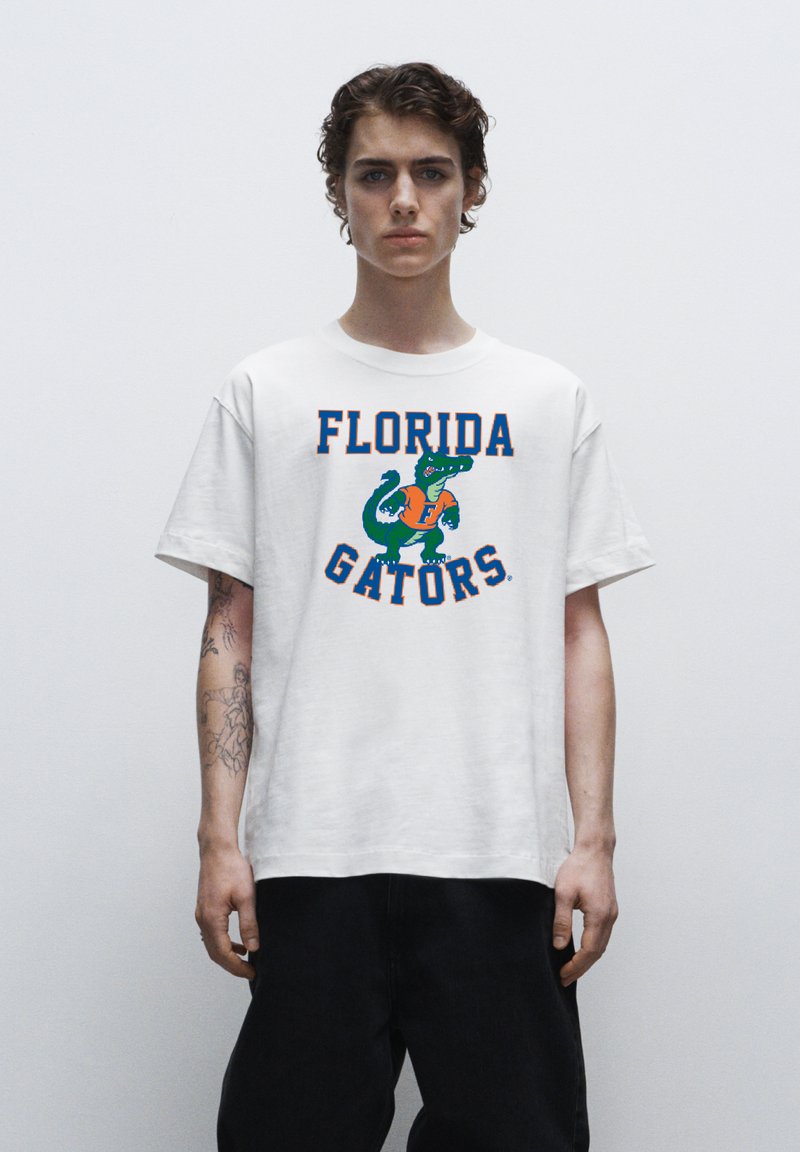 Witte katoenen t-shirt met de tekst "FLORIDA GATORS" in oranje en blauw, met een groene alligator graphic. Ontspannen pasvorm en korte mouwen.