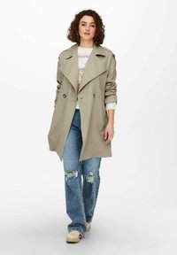 Helle beige Trenchcoat mit breiten Revers und doppelreihiger Knopfleiste. Kombiniert mit ausgebleichten blauen Denim-Jeans und beigen Schuhen.