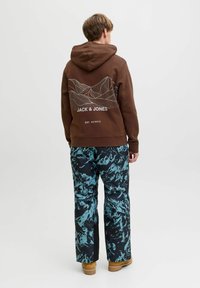 Haut à capuche marron avec un graphisme de ligne de montagne blanche et du texte au dos, associé à un pantalon aux motifs abstraits bleus et noirs, et à des chaussures beiges.