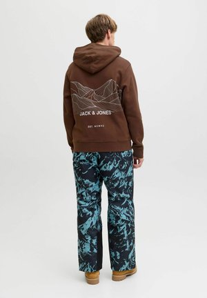 Haut à capuche marron avec un graphisme de ligne de montagne blanche et du texte au dos, associé à un pantalon aux motifs abstraits bleus et noirs, et à des chaussures beiges.
