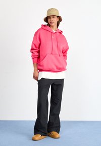 Lys rosa hettegenser med kengurulomme og snor, lagd over en hvit skjorte, kombinert med løse svarte bukser og beige slip-on sko.