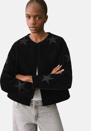 Chaqueta de terciopelo negro con patrones de estrellas, superficie texturizada y un diseño corto. Presenta un cuello redondo y detalles de costura sutiles.