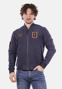 Giacca bomber blu navy con zip, presenta toppe ricamate e polsini a costine, realizzata in un tessuto liscio. Include tasche frontali.