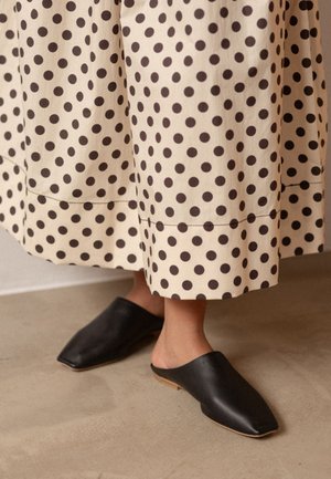 Juodos odinės mūlos su smailiu nosiu, derinamos su kremine iki kelių ilgio suknele, išmarginta dideliais rudais taškeliais.