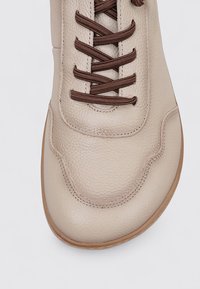 Beige leather shoe with brown laces, rounded toe, textured surface, and light stitching details. Smooth sole with subtle curvature.