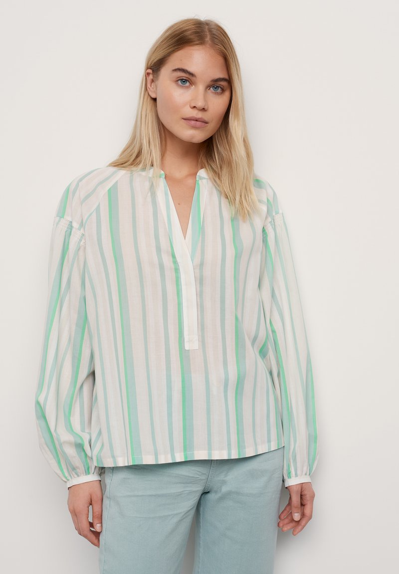 Jeune femme à cheveux blonds longs, portant un chemisier ample à rayures vertes, blanches et beiges, associé à un pantalon bleu clair, se tenant devant un mur blanc.